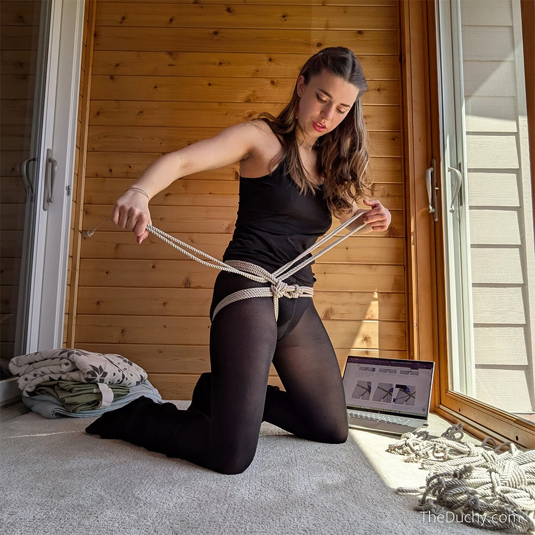 A woman wearing black tights kneels on the floor of a sunlit room. She is pulling rope through a harness she is tying around her hips