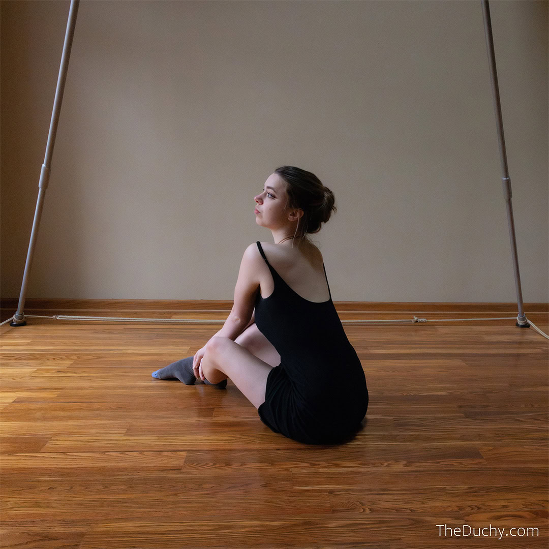 A picture of a young woman with pale skin and brown hair sits on a wood floor. Her back is to the camera and her body is angled to the left. Natural light hits her face and body.