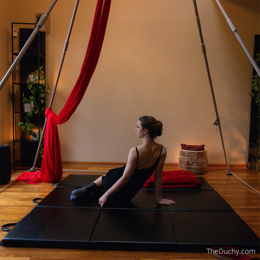 A picture of a young woman with pale skin and brown hair reclining on a black mat. She is in a room with warm light. Houseplants sit on the shelves behind her.