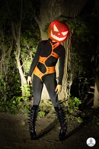 This is a Halloween photograph. Headlights capture a woman with a pumpkin where her head should be. It has a frightening face glowing from within with fiery light. She is wearing a black skin tight catsuit, black patent-leather platform boots and a body harness of orange rope that snakes its way from her throat and runs between her legs, lines run from it around her body to hug her curves.